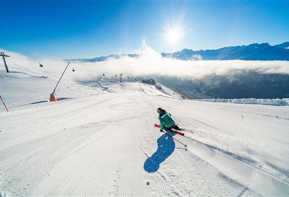 Ein Skifahrer fährt eine perfekt präparierte Piste hinunter. Im Hintergrund sind Berge und ein strahlend blauer Himmel zu sehen.