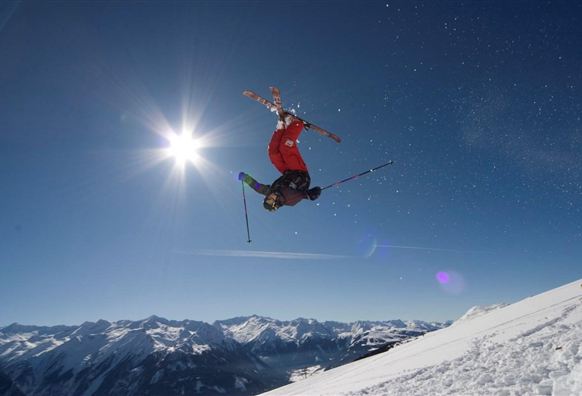 Ein Skifahrer führt einen Sprung in der Luft aus. Der Himmel ist klar und die schneebedeckten Berge sind im Hintergrund sichtbar.