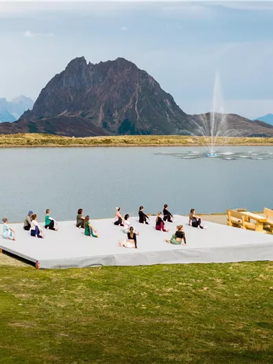 Eine Gruppe von Menschen macht Yoga auf einer Bühne am Ufer eines Sees. Im Hintergrund sind majestätische Berge und ein Wasserstrahl zu sehen.