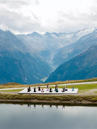 Eine Gruppe von Menschen macht Yoga auf einer Plattform in den Bergen. Im Hintergrund sind beeindruckende Berge und ein klarer See zu sehen.