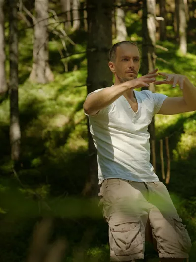 Two people are doing yoga by the shore of a lake. In the background, mountains and boats are visible.