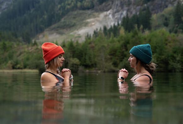 Zwei Frauen stehen im Wasser und schauen sich an. Sie tragen bunte Mützen und die Umgebung ist von grünen Bäumen und Bergen geprägt.