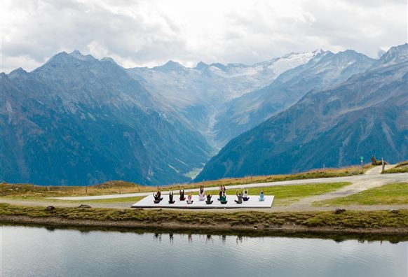 Eine Gruppe von Menschen macht Yoga auf einer Plattform in den Bergen. Im Hintergrund sind beeindruckende Berge und ein klarer See zu sehen.