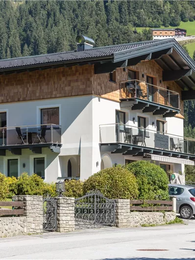 Een modern huis in alpine stijl met een balkon en een verzorgde tuin. Op de achtergrond zijn groene heuvels en bomen te zien.