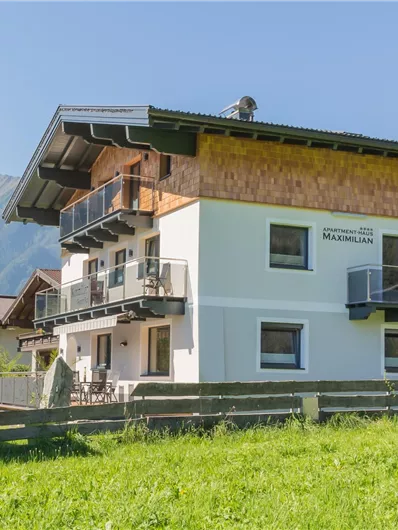 Een modern huis met twee verdiepingen in een landelijke omgeving. Op de achtergrond zijn bergen en een blauwe lucht te zien.