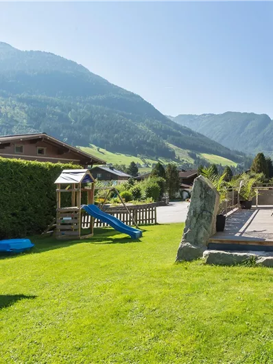 Een mooie tuin met groen gras en speeltoestellen. Op de achtergrond zijn bergen en een blauwe lucht te zien.