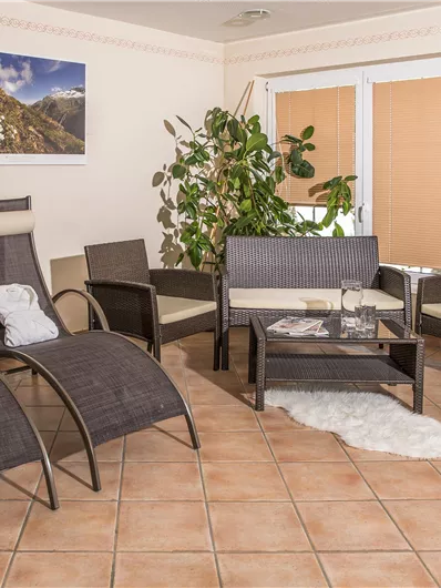 Ein gemütlicher Raum mit modernen Möbeln und einer großen Pflanze. Entspannte Liegen und ein Couchtisch sorgen für eine einladende Atmosphäre.