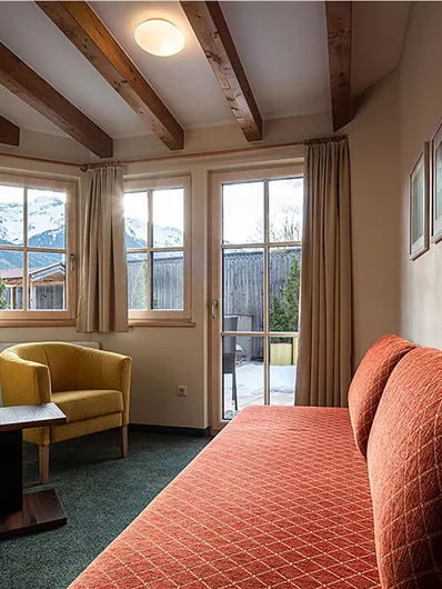 Ein gemütliches Zimmer mit einem orangefarbenen Sofa und einem gelben Sessel. Große Fenster bieten Blick auf die umliegende Natur.