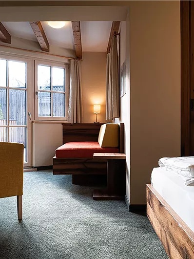 Ein gemütliches Zimmer mit einem Doppelbett, einem Sessel und einer Sitzecke. Große Fenster sorgen für helles Licht und Blick nach draußen.