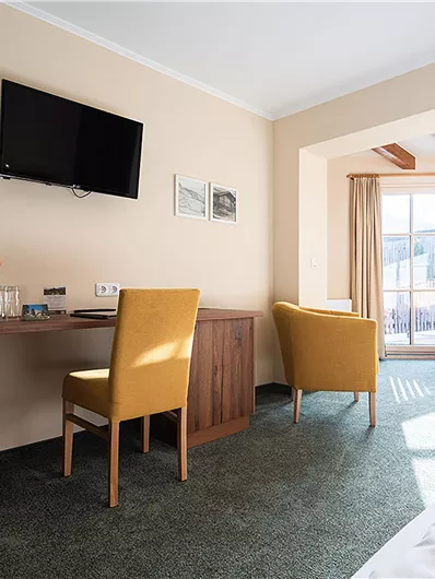 Ein helles Zimmer mit einem Schreibtisch, einem Fernseher und zwei Stühlen. Große Fenster sorgen für viel Tageslicht.