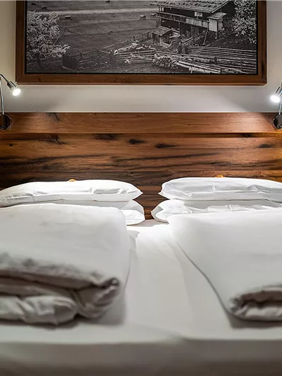 Ein gemütliches Doppelbett mit weißen Bettdecken und einer hölzernen Kopfteil. An den Seiten befinden sich Nachtlichter und ein Bild an der Wand.