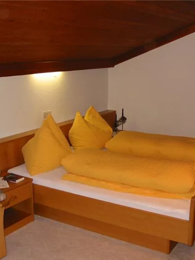 Een gezellige zolderkamer met een groot bed en gele kussens. In de hoek staat een comfortabele stoel en een kleine plantendecoratie.