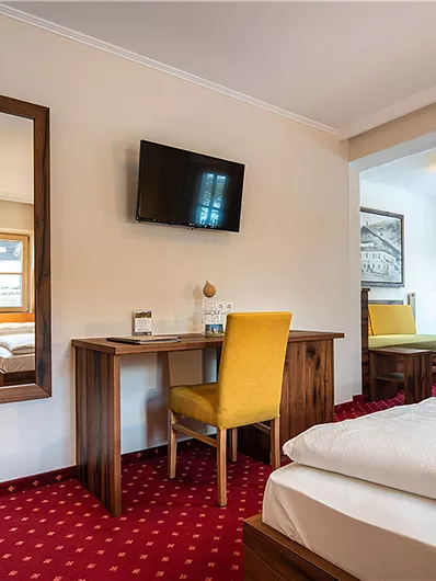 Ein helles Hotelzimmer mit einem Doppelbett, einem Tisch und einem Fernseher. Im Hintergrund sind ein großer Spiegel und ein Stuhl zu sehen.