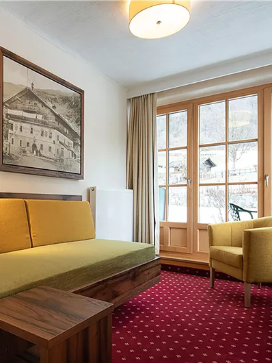 Ein gemütliches Zimmer mit einem grünen Sofa und einem gelben Sessel. Große Fenster lassen viel Licht herein und bieten einen Blick nach draußen.