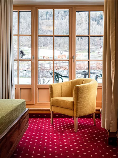 Ein gemütliches Zimmer mit einem gelben Sessel und einem roten Teppich. Große Fenster bieten einen Blick nach draußen auf die Landschaft.