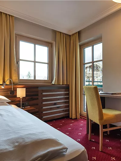 Ein gemütliches Hotelzimmer mit einem Einzelbett, einem Schreibtisch und einem Fernseher. Große Fenster lassen viel Tageslicht herein und bieten einen Blick nach draußen.