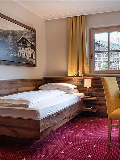 Ein gemütliches Hotelzimmer mit einem Einzelbett, einem Schreibtisch und einem Fenster mit Aussicht. Die Wände sind in warmen Holzfarben gehalten und die Vorhänge sind goldgelb.