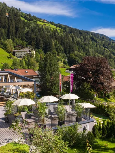 Ein malerisches Hotel mit einer Terrasse, umgeben von grünen Bergen. Die Sonnenschirme laden zum Entspannen im Freien ein.