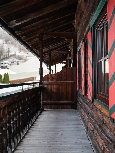 Ein Holzveranda mit rot-grünen Fensterläden. Im Hintergrund sind schneebedeckte Hügel und ein kleines Dorf zu sehen.