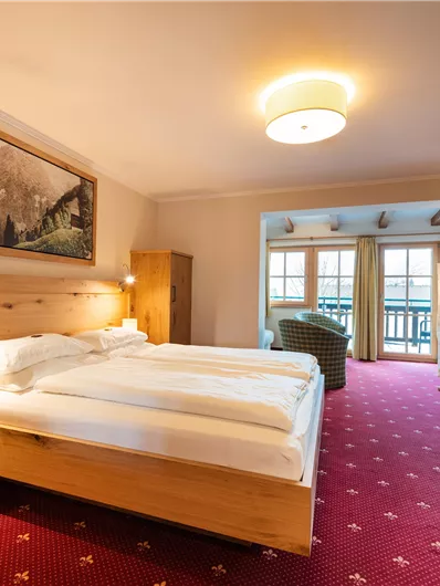 Ein modernes Hotelzimmer mit einem Doppelbett und einem gemütlichen Sessel. Große Fenster bieten Zugang zu einem Balkon mit Blick nach draußen.