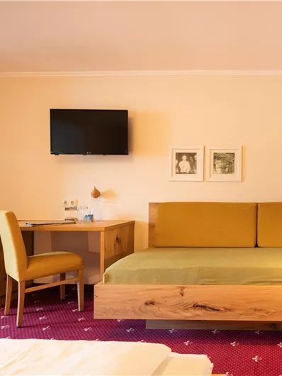 Ein gemütliches Zimmer mit einem Holzbett, einem Schreibtisch und einem Fernseher. Die Einrichtung ist hell und freundlich mit einem roten Teppich.