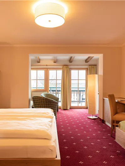 Ein gemütliches Hotelzimmer mit einem Doppelbett, einem Tisch und einem Sessel. Große Fenster bieten einen Blick ins Freie und lassen viel Licht herein.