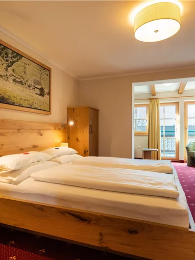 Ein gemütliches Hotelzimmer mit zwei Betten aus Holz. Es gibt einen Schreibtisch, einen Sessel und Fenster mit Vorhängen.