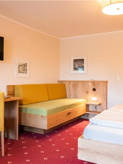Ein gemütliches Hotelzimmer mit zwei Betten und einem kleinen Tisch. An der Wand hängt ein Fernseher und es gibt eine gelbe Sitzecke.