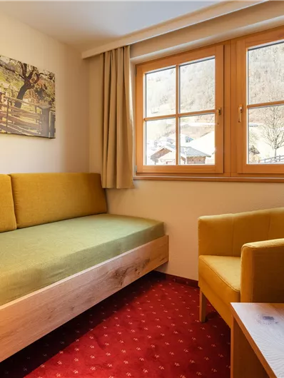 Ein gemütlicher Aufenthaltsraum mit einem grünen Sofa und einem gelben Sessel. Große Fenster lassen viel Licht herein und bieten einen Blick nach draußen.