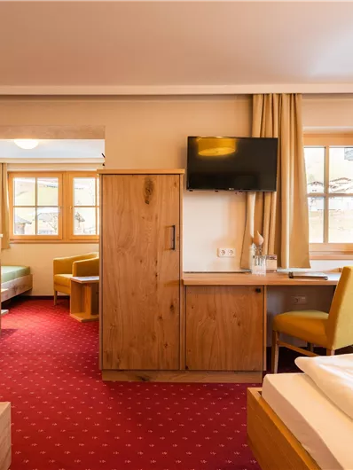 Ein gemütliches Zimmer mit Holzmöbeln und roten Teppichboden. Es gibt einen Fernseher, einen Schreibtisch und ein bequemes Sofa.