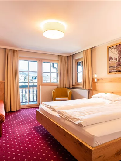 Ein gemütliches Hotelzimmer mit einem Doppelbett und einem Schreibtisch. Das Zimmer hat große Fenster und einen Balkon mit Blick nach draußen.