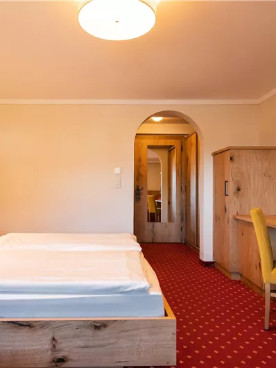 Ein gemütliches Hotelzimmer mit zwei Betten und einem Schreibtisch. Der Raum ist hell und einladend, mit rotem Teppichboden und einem Fernseher an der Wand.