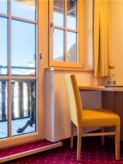 Ein gemütliches Zimmer mit einem Schreibtisch und einem Stuhl. Der Blick führt auf einen Balkon mit Aussicht auf die Berge.
