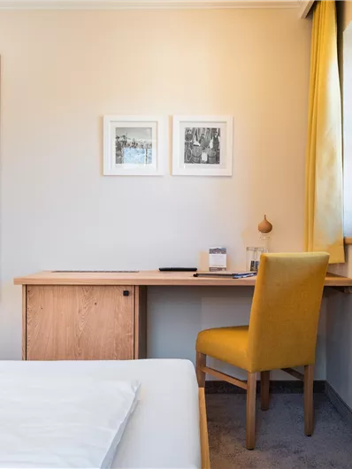 Ein modernes Hotelzimmer mit einem Doppelbett, einem Schreibtisch und einem gelben Stuhl. An der Wand hängen Bilder und ein Spiegel reflektiert das Licht.