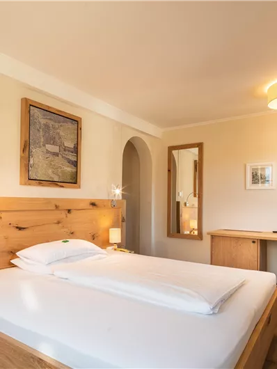 Ein gemütliches Schlafzimmer mit einem Doppelbett und warmen Holzmöbeln. Die Wände sind hell gestrichen und es gibt einen Schreibtisch sowie dekorative Kunstwerke.