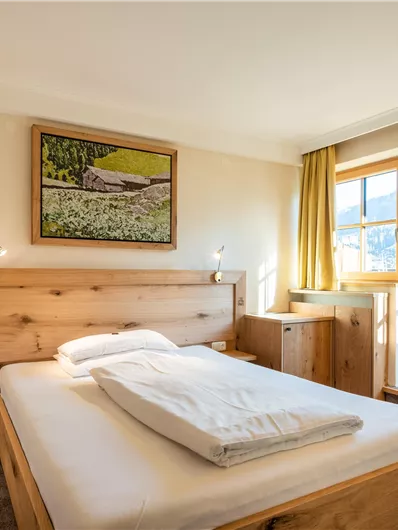 Ein gemütliches Zimmer mit einem Doppelbett und hellen Holzdetails. Große Fenster mit gelben Vorhängen sorgen für viel Tageslicht.