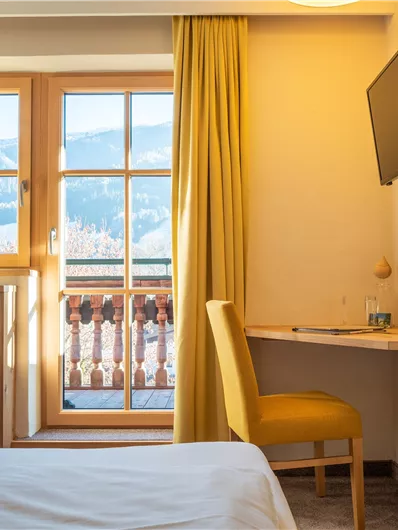 Ein gemütliches Zimmer mit einem großen Fenster und gelben Vorhängen. Der Raum ist modern eingerichtet mit einem Schreibtisch und einem Fernseher.