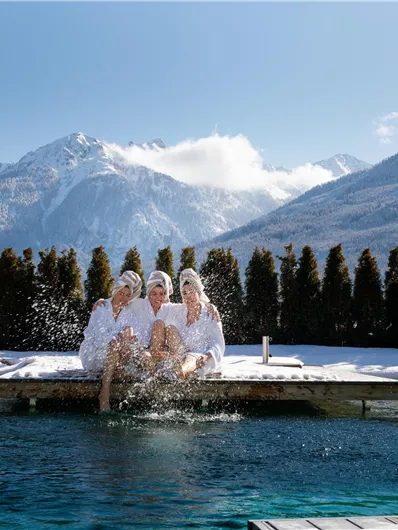 Drie vrouwen in badjassen zitten aan een zwembad in een winterachtig berglandschap. De lucht is helder en de bergen zijn bedekt met sneeuw.