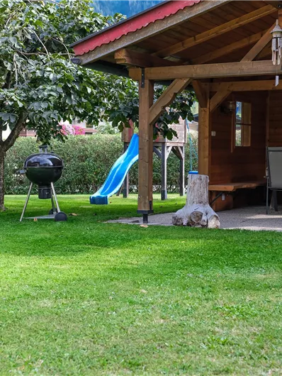 A cozy garden with a wooden terrace and a grill. In the background, there is a playset and a lawn.