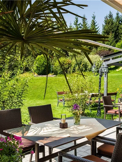 Ein schöner Außenbereich mit Tischen und Stühlen umgeben von Pflanzen. Die grüne Wiese und Bäume bieten eine entspannte Atmosphäre.