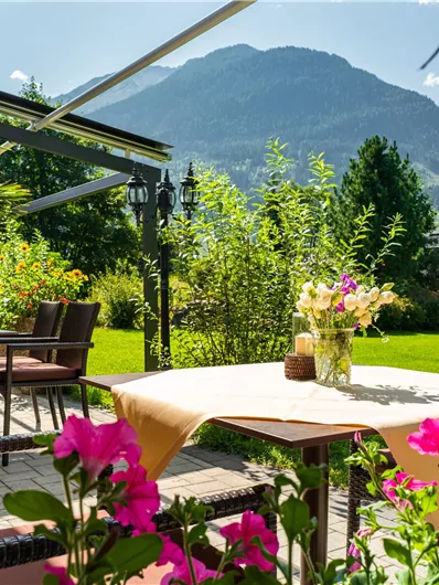 Eine einladende Terrasse mit Tischen und bunten Blüten. Im Hintergrund erheben sich majestätische Berge unter einem klaren Himmel.