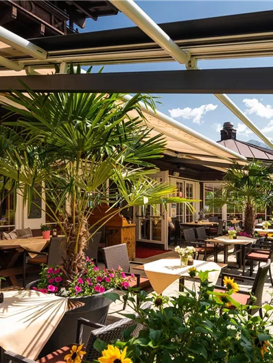 Eine gemütliche Terrasse mit Tischen und Stühlen, umgeben von Pflanzen und Blumen. Im Hintergrund sind Berglandschaften und ein blauer Himmel zu sehen.