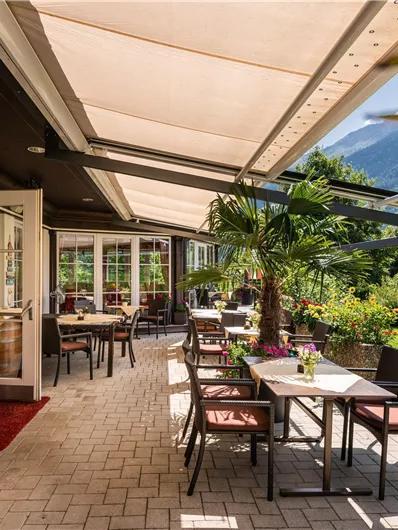 Ein einladender Außenbereich eines Restaurants mit Tischen und Stühlen. Im Hintergrund sieht man Berge und eine grüne Landschaft.