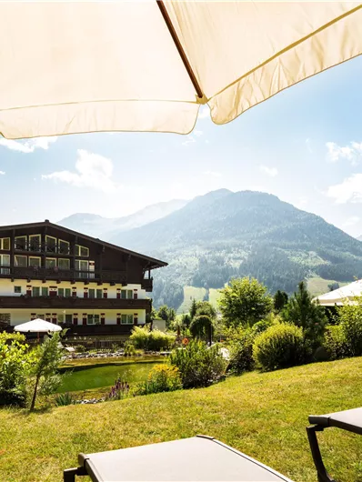 Ein gemütliches Hotel in einer schönen Berglandschaft. Die Umgebung ist grün, mit Sonnenliegen und Sonnenschirmen.