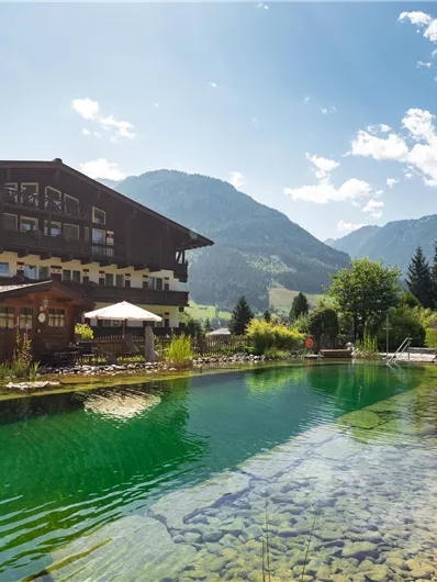 Ein malerisches Hotel am Ufer eines klaren Teichs, umgeben von grünen Wiesen und Bergen. Die Sonne scheint und der Himmel ist blau mit ein paar Wolken.