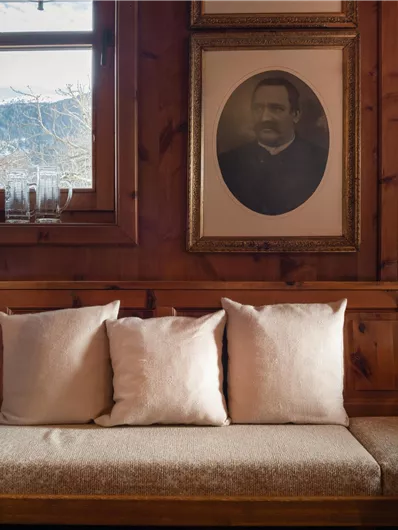 Ein gemütlicher Holzraum mit einer Bank und Kissen. An der Wand hängt ein Portrait und große Fenster bieten einen Blick auf die Berge.