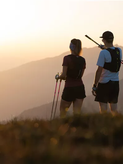 Two hikers stand on a hill and look at the mountains in the sunset. They hold trekking poles in their hands and wear sports clothing.