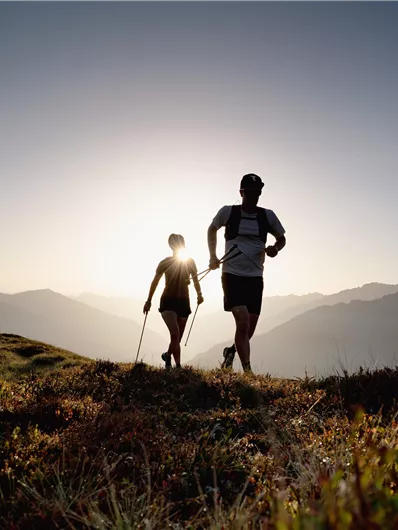 Two people are jogging in nature with mountains in the background. The sun is shining and creates a romantic atmosphere.