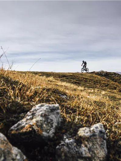 A cyclist rides over a hill with rocky terrain. The sky is cloudy and the landscape is plain and natural.