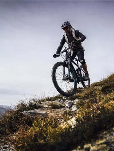A mountain biker rides on a narrow path in a mountainous landscape. The surroundings are characterized by green and gray tones, with a cloudy sky in the background.
